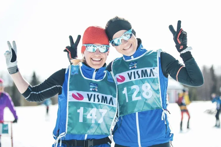Kaksi hymyilevää hiihtäjää kilpailunumeroissa näyttää voitonmerkkiä