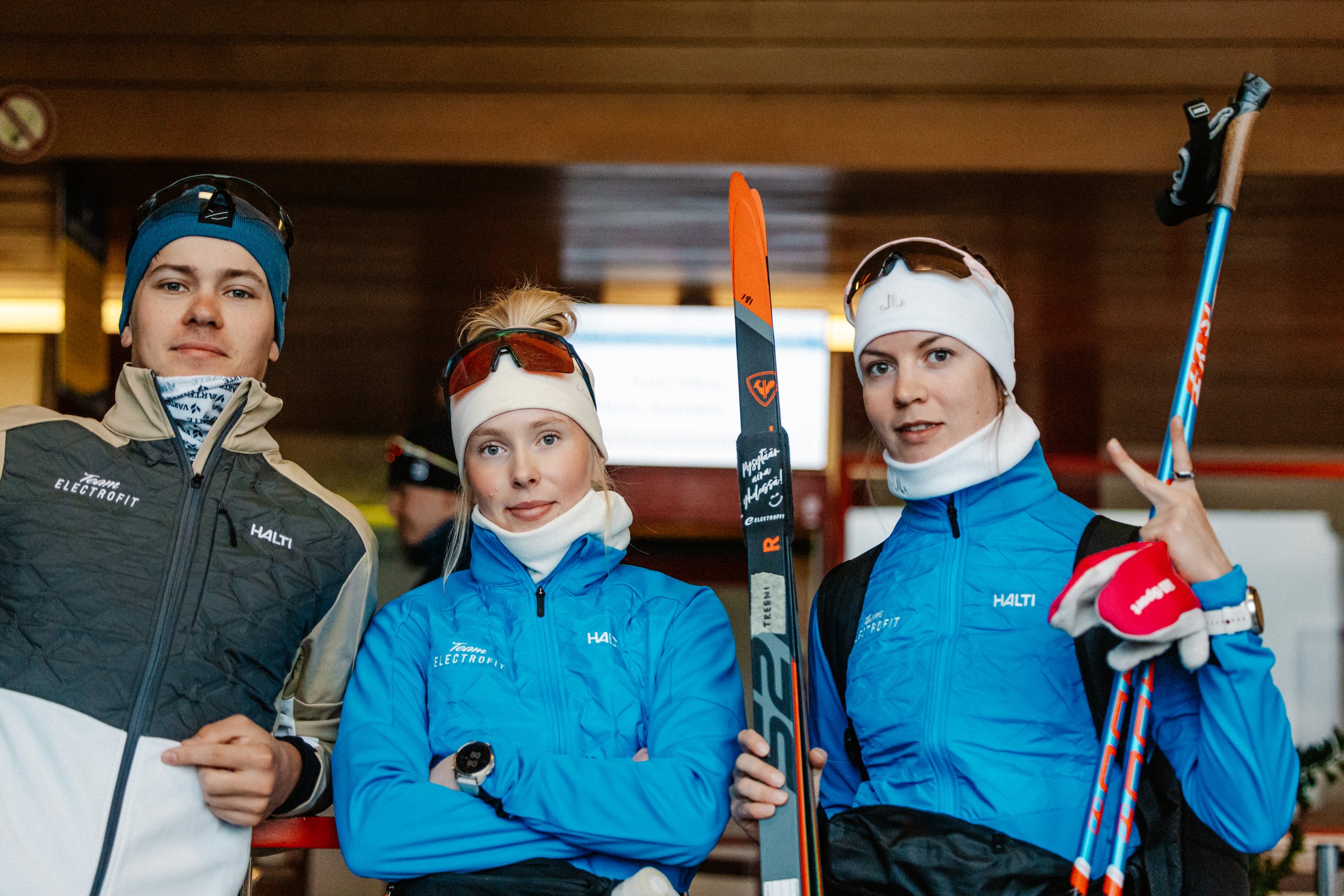 Kolme Team Electrofitin hiihtäjää suksineen ja sauvoineen hymyilee kameralle sisätiloissa.