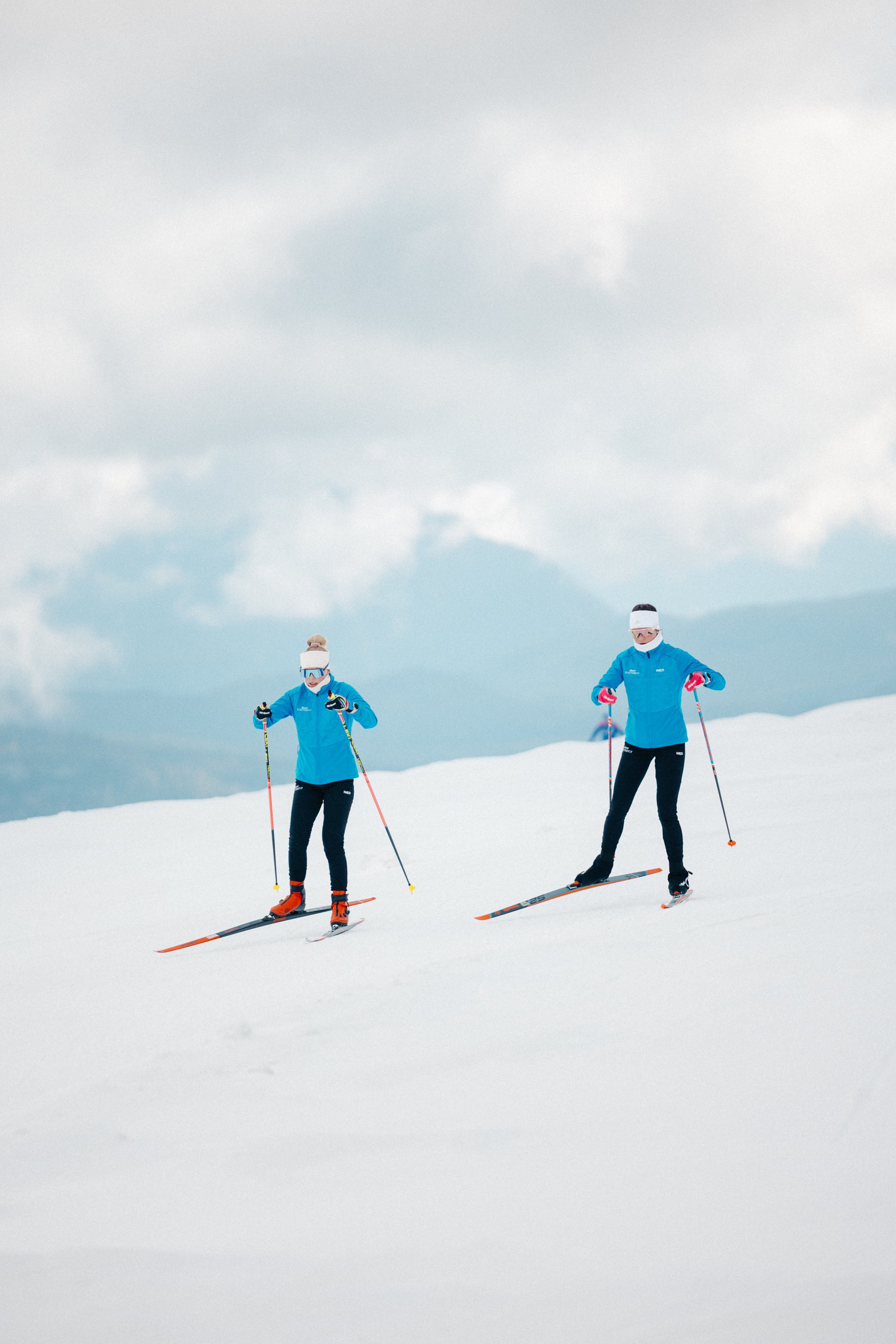 Kaksi hiihtäjää sinisissä Team Electrofitin takeissa hiihtää lumisella jäätiköllä, taustalla pilvien peittämät vuoret.